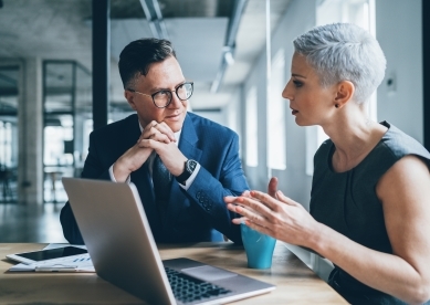 Business man in glasses listening to business woman talk in meeting room with laptop in front of them